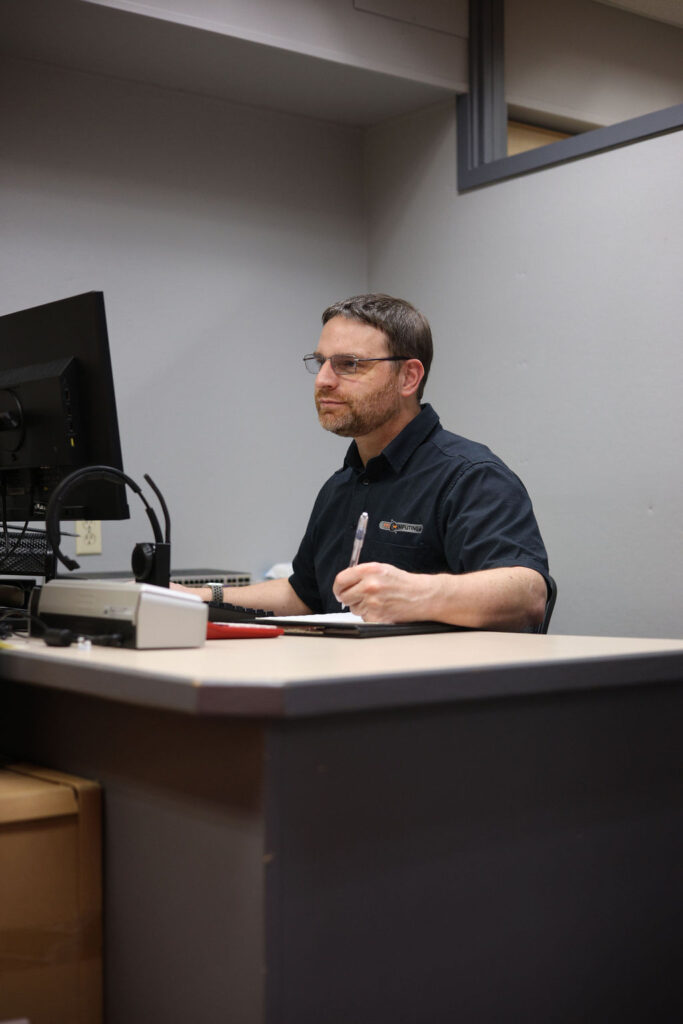 A man sits at his desk with a pen in hand ready to write on a piece of paper while he looks at the computer screen with a soft smile. Glasses rest on his face and he is wearing a branded Faster Than Light Computing black shirt. There are a few electronics in the foreground of the picture and a computer screen partially in view