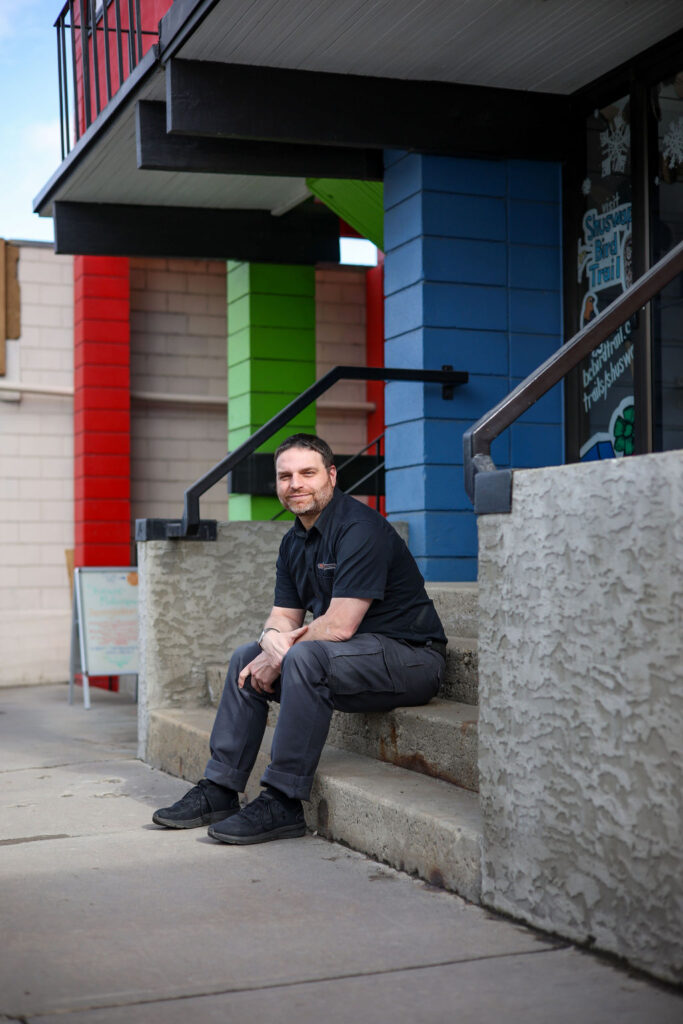 Man sits on the second step of a 4 step staircase leading into a colourful building called the Innovation Centre. His arms are corssed gently as the sun hiits his face and he smiles at the camera. His eyes are slightly squinting due to the sunshine and a sign welcoming people to the Shuswap Makerspace is blurred out behind him.  He wears a branded Faster Than Light Computing black shirt with grey pants that have cargo pockets and black running shoes.