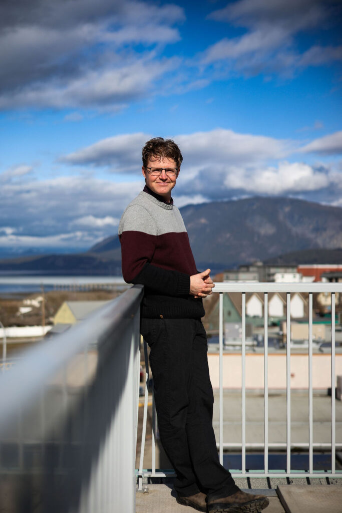 Man stand on a roof top deck in a burgandy and grey wool sweater with black straight leg pants. He is leaning on a metal railing. Shuswap Lake and Bastion mountain of Salmon Arm are in the distance behind him. The sky is moody with clouds but also bright blue peeking out.