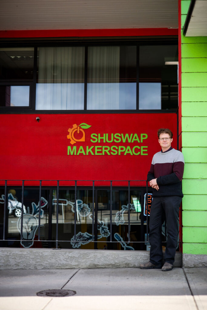 Man stands outside a building with the words Shuswap Maker space on the side of the building  in green. The building is bright multi colored with big windows that have flowers drawn on the glass. The man is standing with his  hand lightly crossed and a small smile on his face. He is wearing a burgundy and grey wool sweater and a pair of black pants with brown shoes. 