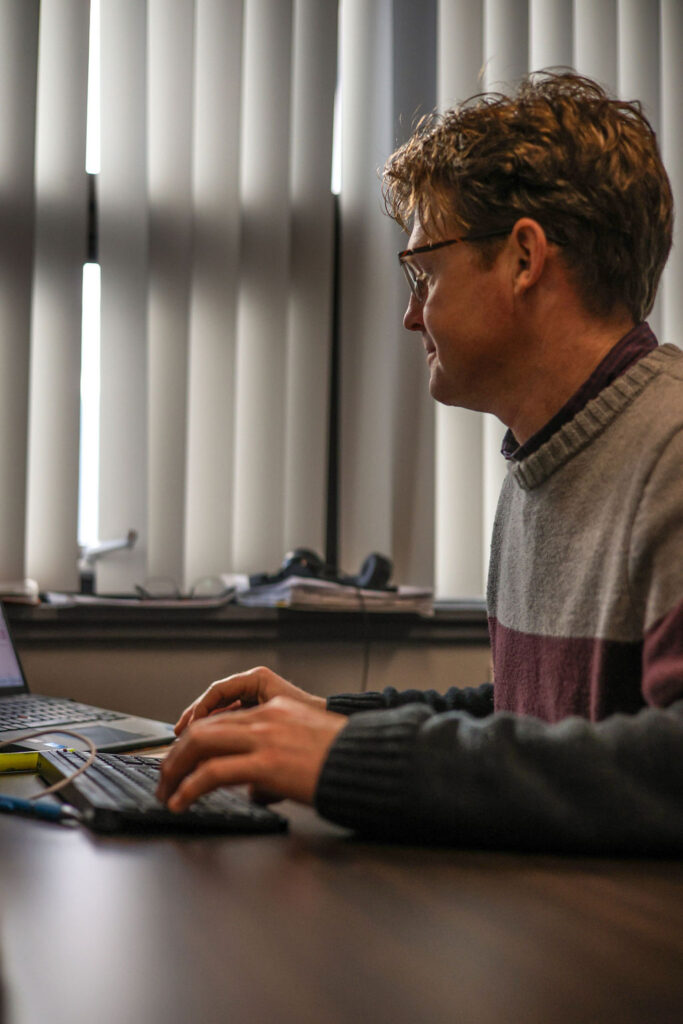  A man sits at his desk looking at his computer and typing on a keyboard. He is wearing a burgundy and grey wool sweater. He has a small smile a pair of glasses resting on his face. Blinds cover the window behind him so there is little natural light in the space.