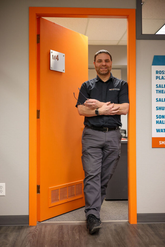 Man standing in an orange door with the letters M4 on it. He is in the doorway wearing branded Faster Than Light Computing, black shirt and grey pants with cargo pockets. His arms are crosses as he hold glasses and smiles at the camera.